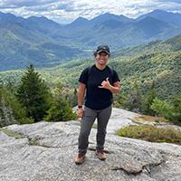 alexis rocio guillermo standing on rocks wearing a black tshirt and grey pants. The background show mountain ranges